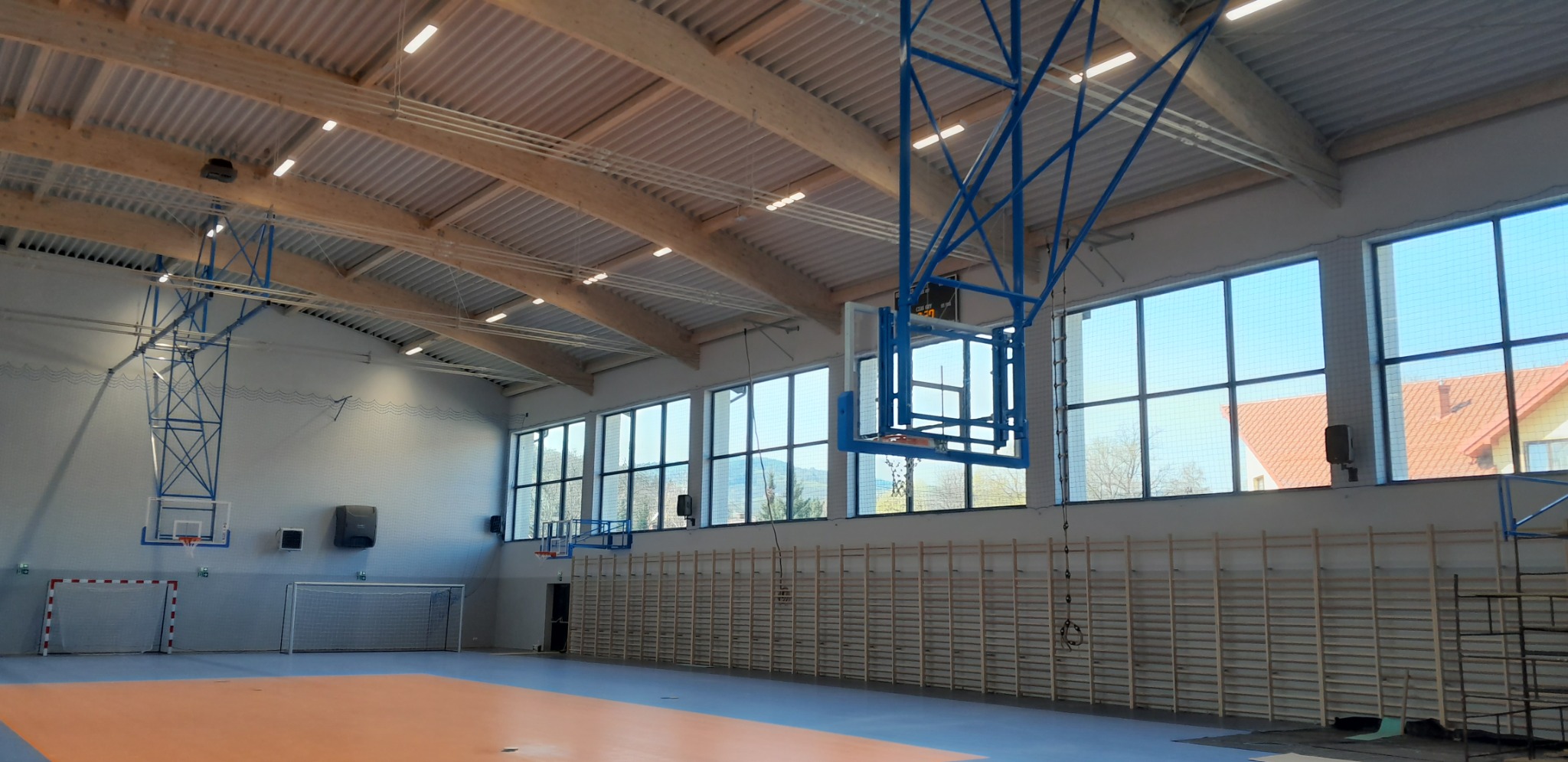 Wnętrze hali sportowej z widocznymi elementami wyposażenia: kosze do koszykówki, bramki do piłki ręcznej, drabinki gimnastyczne wzdłuż ściany oraz zainstalowane głośniki na ścianach.