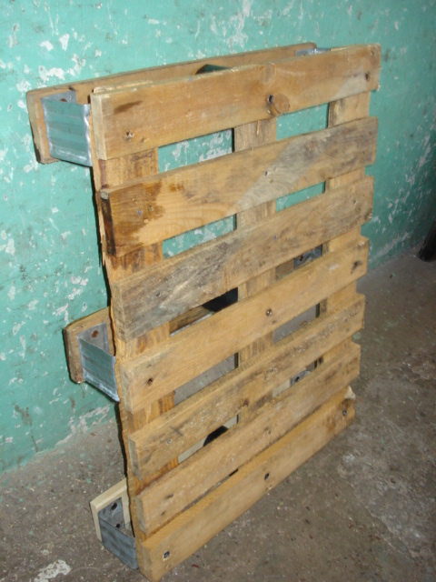 Używana, drewniana paleta z metalowymi wzmocnieniami narożników, oparta o ścianę w pomieszczeniu.