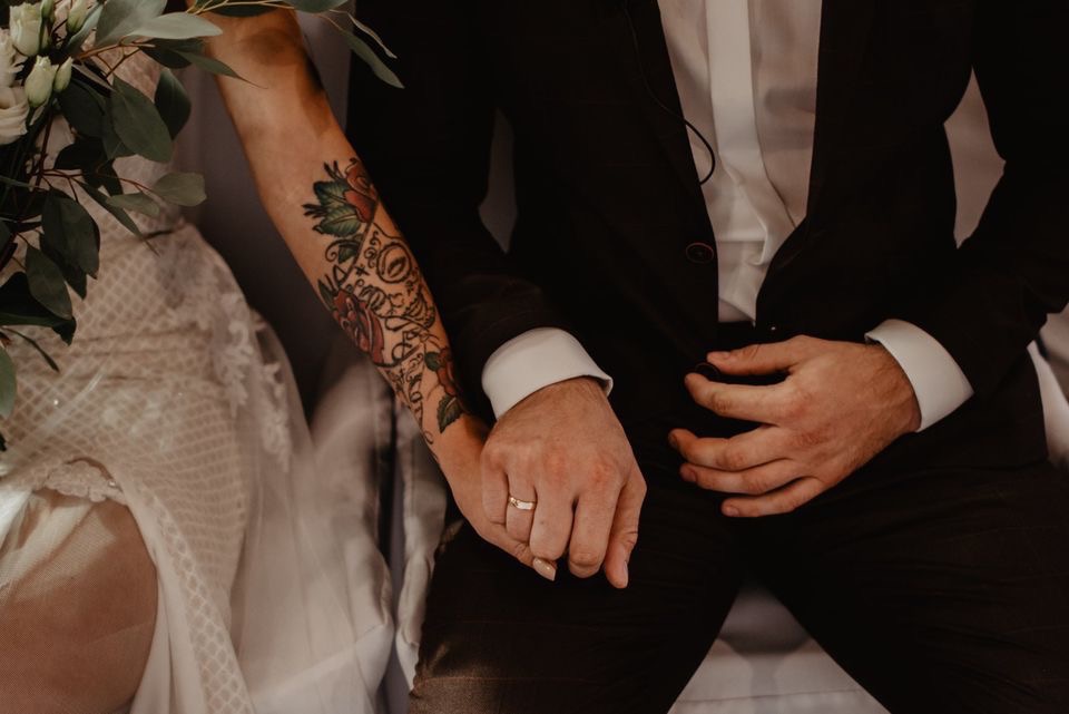 Para młoda z Gryfic trzyma się za ręce; panna młoda w sukni z koronką, pan młody w czarnym garniturze, widoczny tatuaż na ramieniu panny młodej i obrączka.