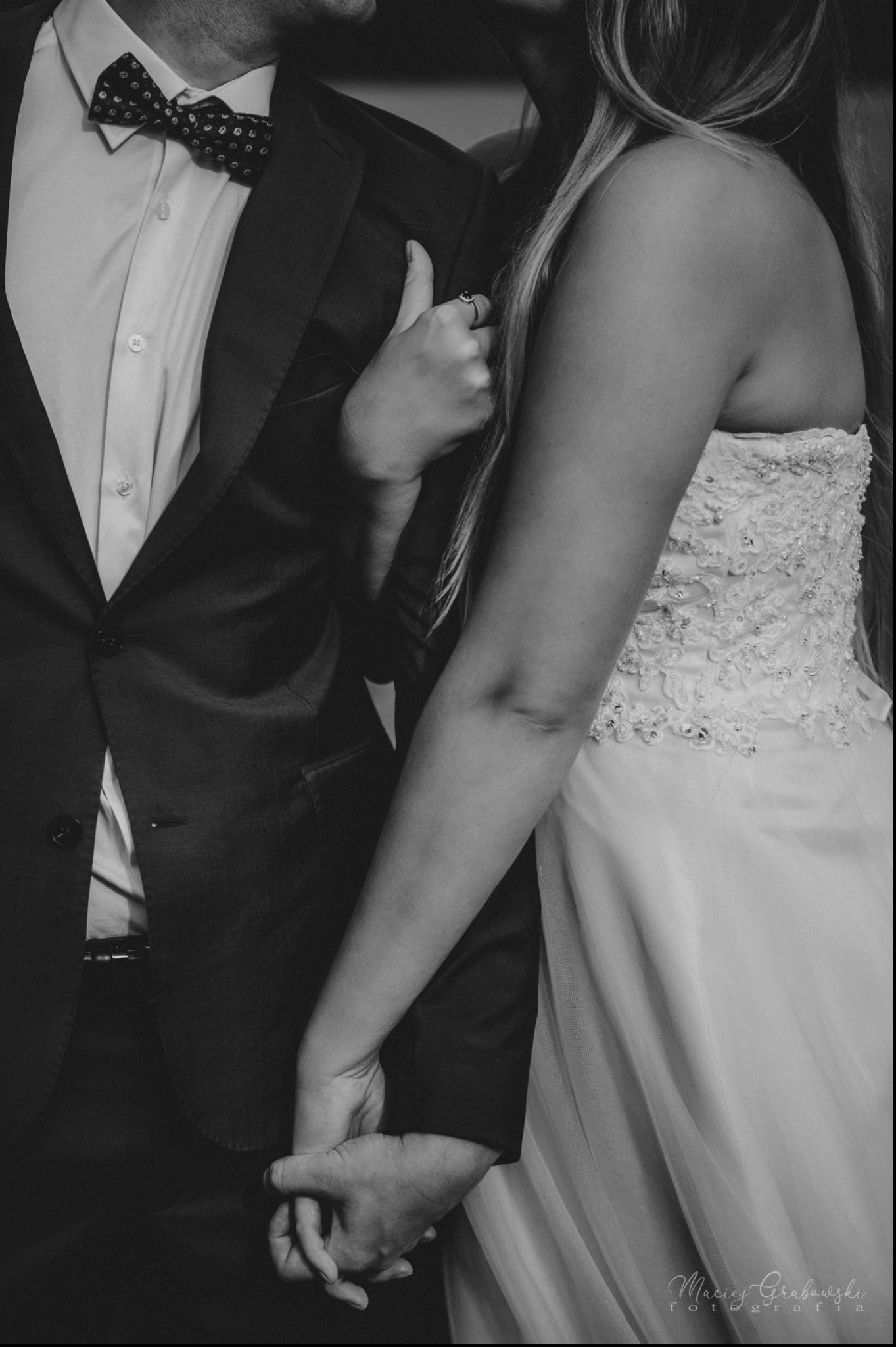 Czarno-białe zbliżenie na parę trzymającą się za ręce: on w ciemnym garniturze z muchą w kropki, ona w sukni ślubnej z koronkową górą i pierścionkiem zaręczynowym.