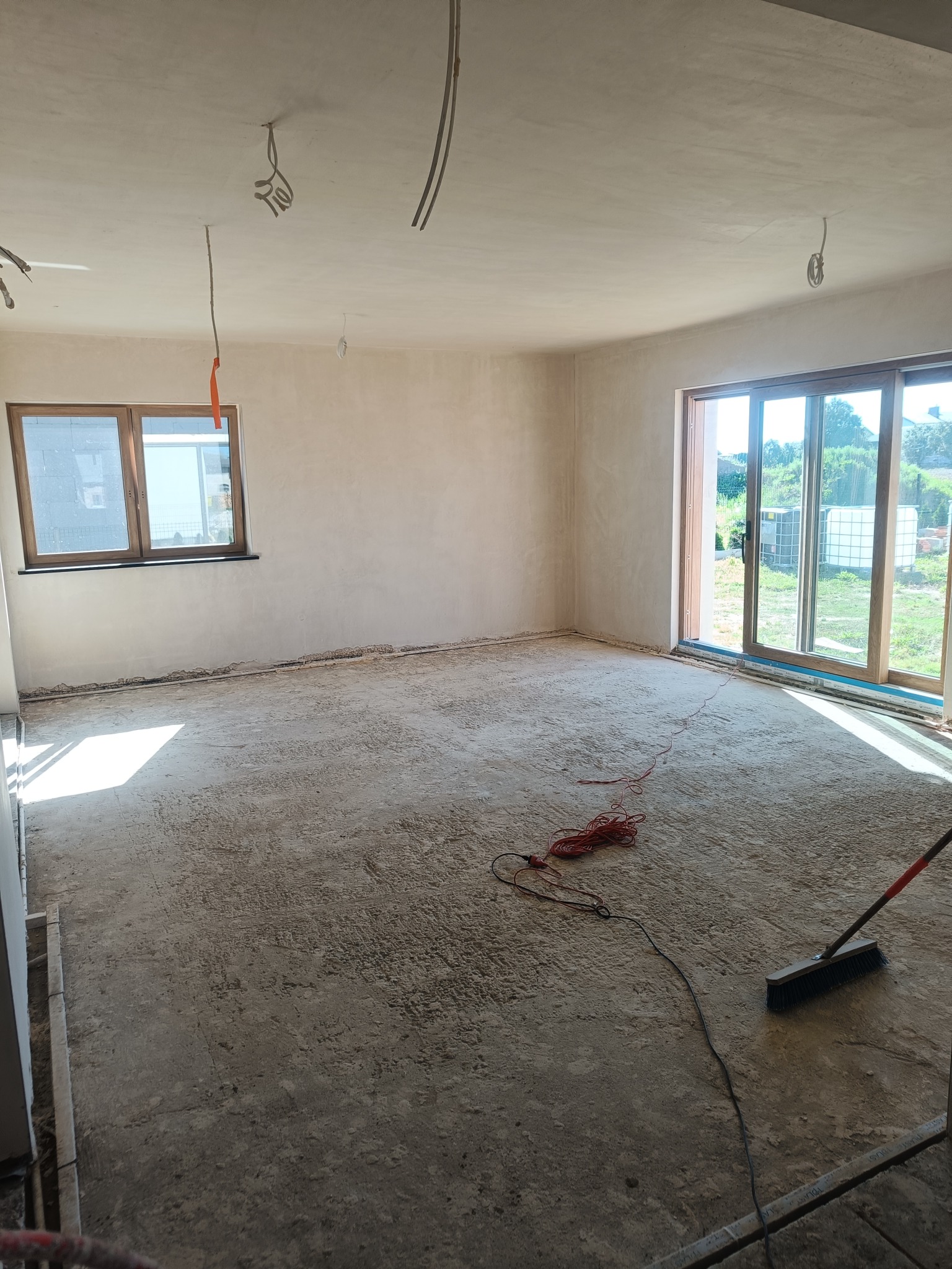 Pokój po nałożeniu tynków: ściany gładkie, surowe podłoże, widoczne przewody elektryczne i narzędzia do sprzątania.