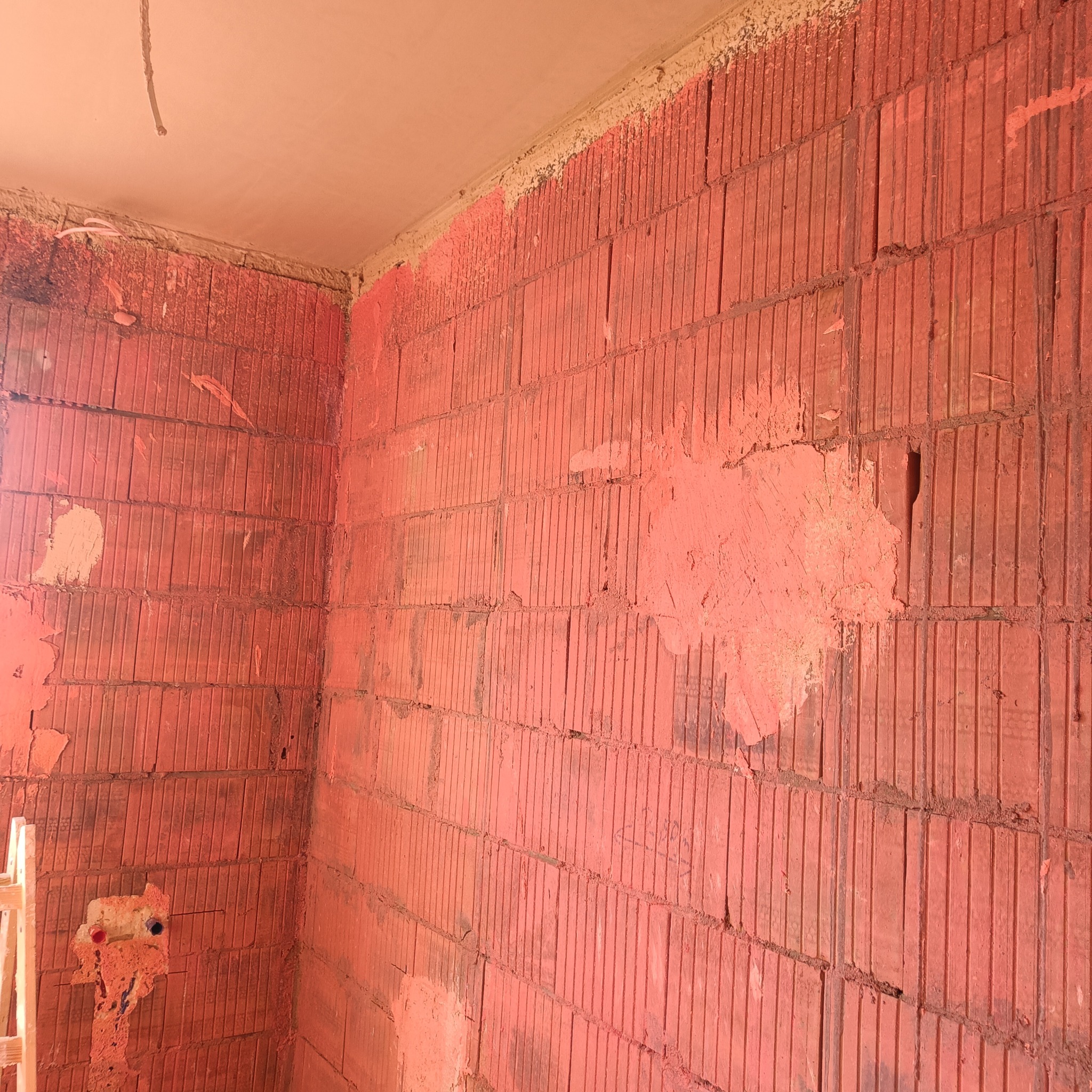 Czerwona ściana z pustaków ceramicznych w trakcie prac tynkarskich, widoczne fragmenty surowego muru i nałożonego tynku w odcieniach różu, częściowo zakryte instalacje wodne, fragment drabiny w rogu.