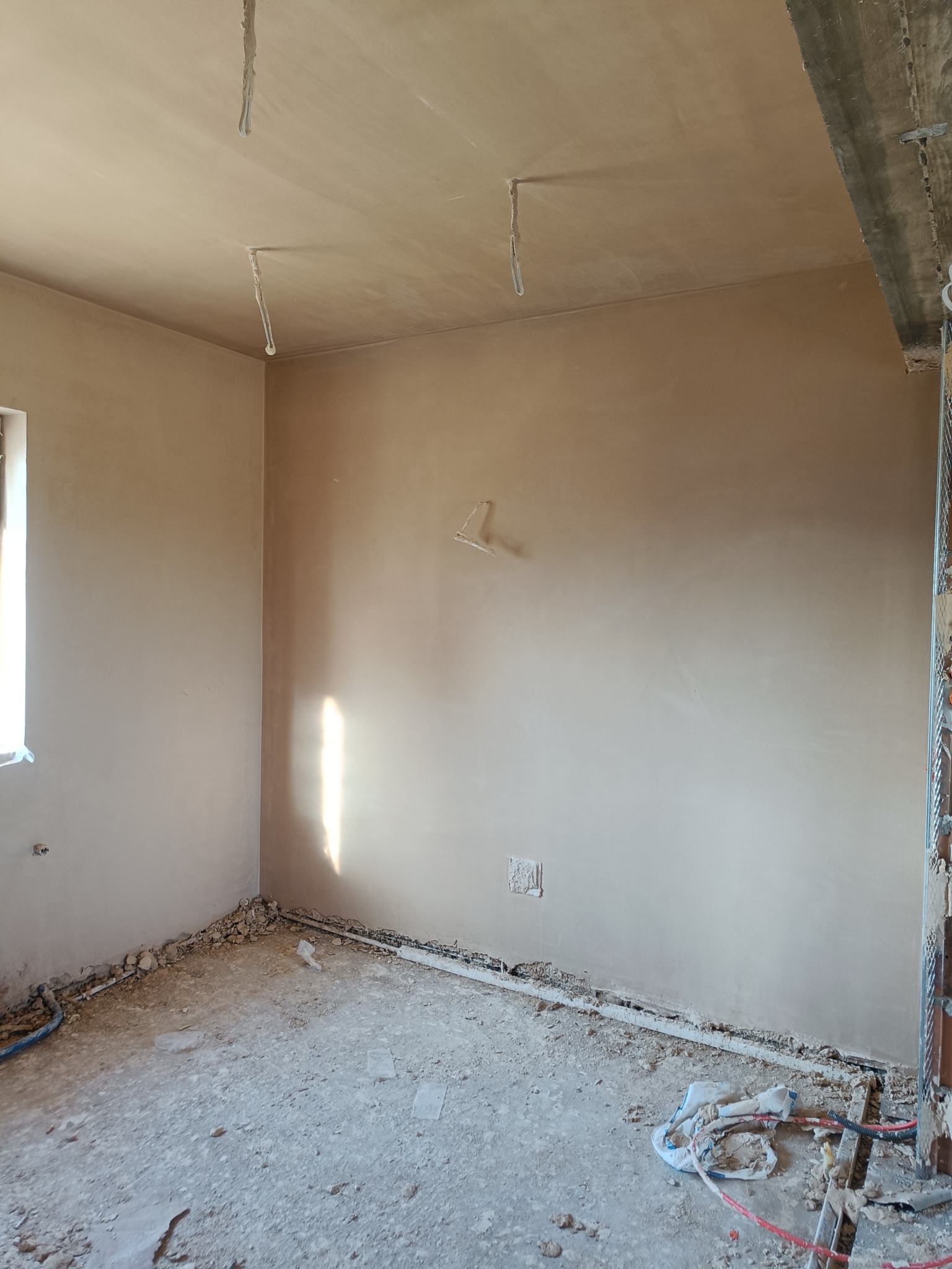 Pokój po nałożeniu tynków maszynowych; widoczne wystające przewody elektryczne ze ściany i sufitu, oraz resztki materiałów budowlanych na podłodze.