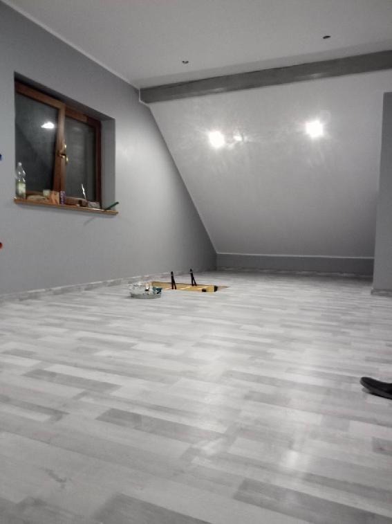 Świeżo otynkowane poddasze ze skośnym sufitem, widoczne narzędzia na podłodze i fragment butelki na parapecie okna.