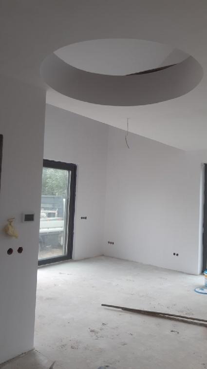 Wnętrze po nałożeniu gładzi, widoczny okrągły element dekoracyjny na suficie, ślady po pracach na podłodze, dwa okna w czarnych ramach.