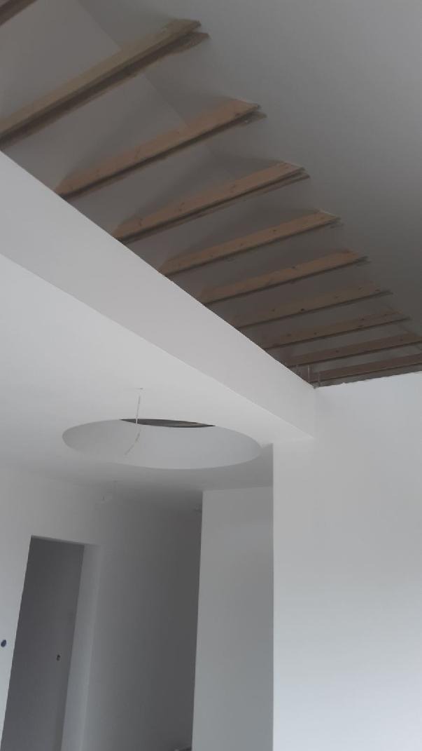 Wnętrze w trakcie wykańczania: surowy strop z drewnianymi belkami, gładkie, białe ściany i okrągły otwór w suficie na oświetlenie.