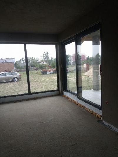 Surowy stan wnętrza z nałożonymi tynkami, widoczne duże okna przesuwne z widokiem na ogród, podłoga przygotowana pod wylewkę.