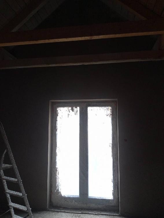 Świeżo nałożony, ciemny tynk na ścianie w pokoju z oknem; widoczna drewniana konstrukcja dachu i drabina oparta o ścianę.