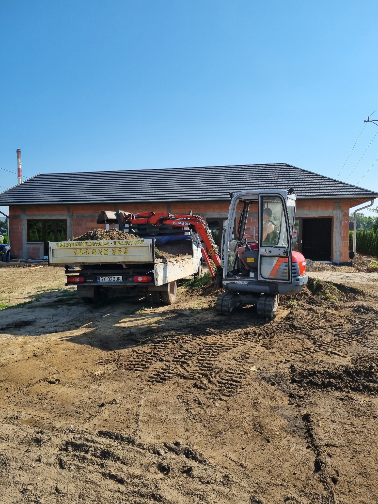 Mini-koparka Kubota ładuje ziemię na wywrotkę na tle budynku w trakcie budowy, widoczne ślady opon na ziemi.