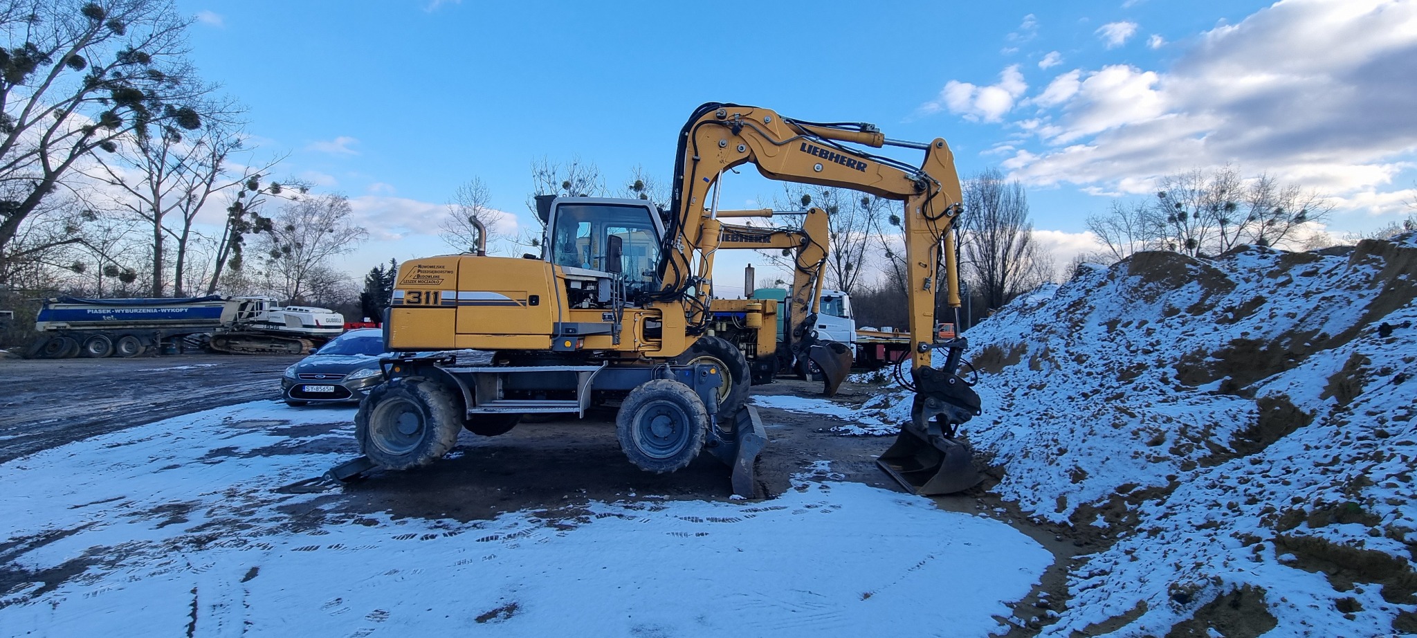Dwa żółte, kołowe koparki Liebherr na placu budowy z niewielką ilością śniegu, widoczny samochód osobowy i ciężarowy w tle, błękitne niebo.