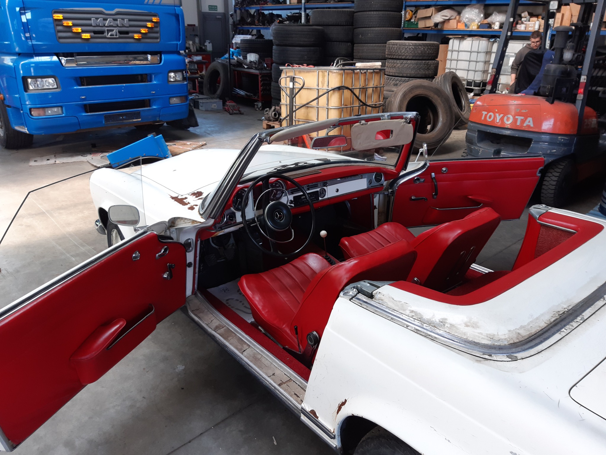 Wnętrze białego, klasycznego kabrioletu z czerwonymi skórzanymi siedzeniami i otwartymi drzwiami, widoczne ślady użytkowania na karoserii, w tle niebieska ciężarówka MAN i wózek widłowy Toyota.
