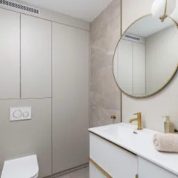 Nowoczesna łazienka w beżach i bieli z akcentami złota: okrągłe lustro, armatura i detale szafki pod umywalką. Na ścianie marmurowe płytki, podwieszana toaleta i zabudowa meblowa.