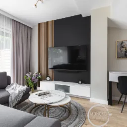 Nowoczesny salon z szarą sofą narożną, marmurowymi stolikami kawowymi, czarną ścianą z drewnianymi lamelami i biurkiem z czarnym krzesłem w rogu