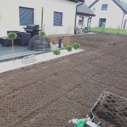 Przygotowane podłoże pod trawnik z rolki, widoczne wyrównane podłoże, obrzeża z białego kamienia, ozdobne krzewy i elementy małej architektury ogrodowej w tle.