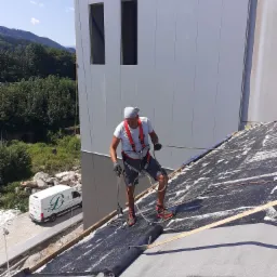 Pracownik w uprzęży zabezpieczającej, stojący na dachu pokrytym czarną membraną, z widoczną rolką membrany i narzędziem do zgrzewania, w tle budynek z szarej blachy i górzysty krajobraz.