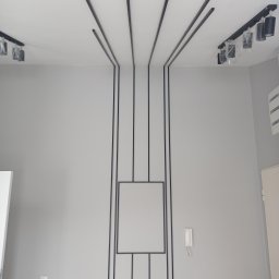 Genpro Pruchenski Grzegorz - Szara ściana z dekoracyjnymi czarnymi listwami tworzącymi geometryczny wzór, oświetlenie sufitowe z czarnymi reflektorami, widoczna skrzynka z instalacją hydrauliczną.