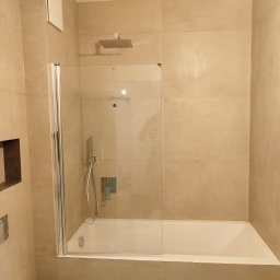 Genpro Pruchenski Grzegorz - Beżowa łazienka z wanną i szklanym parawanem. Widoczna bateria prysznicowa, wnęka w ścianie i chromowany przycisk spłuczki. Minimalistyczny design.