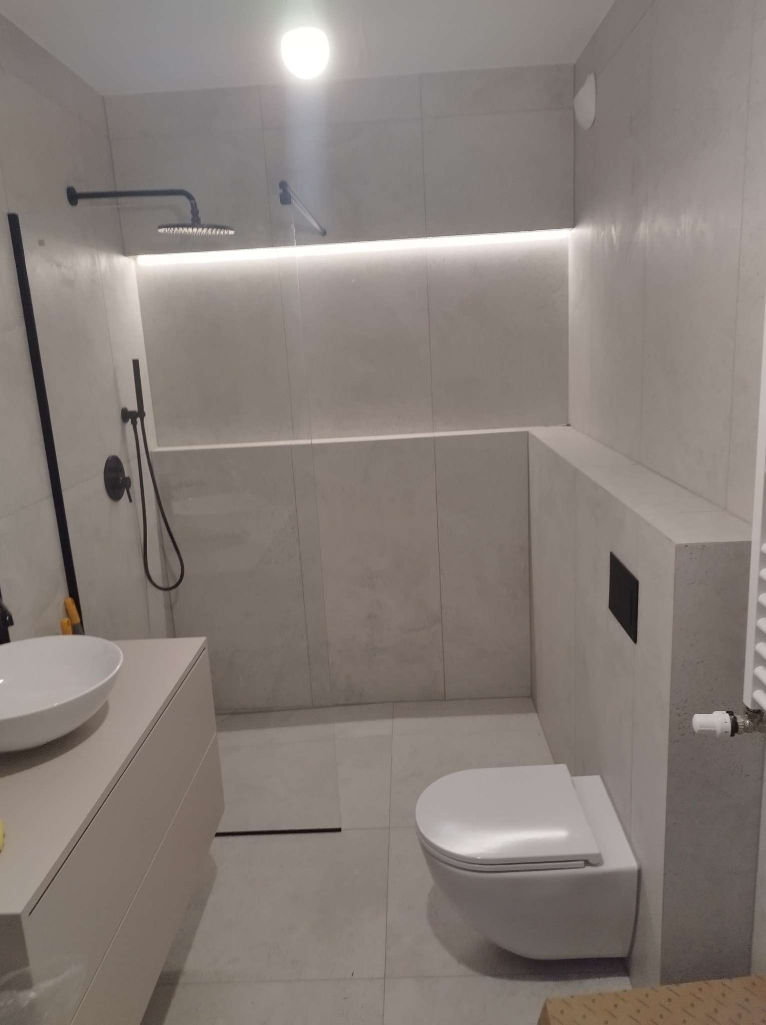 Minimalistyczna łazienka z szarymi płytkami, podwieszaną toaletą, umywalką nablatową i czarną armaturą prysznicową.