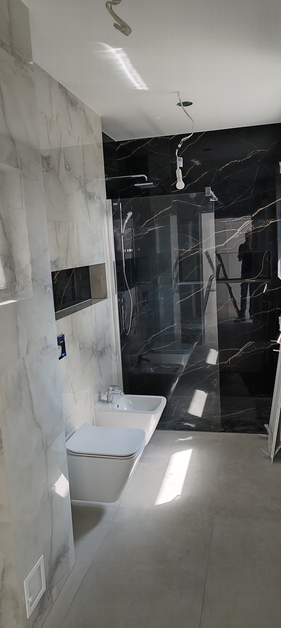 Nowoczesna łazienka w trakcie wykańczania z biało-szarymi marmurowymi płytkami na ścianach, czarną kabiną prysznicową z złotymi żyłami, wiszącą toaletą i bidetem, widoczne niedokończone oświetlenie...
