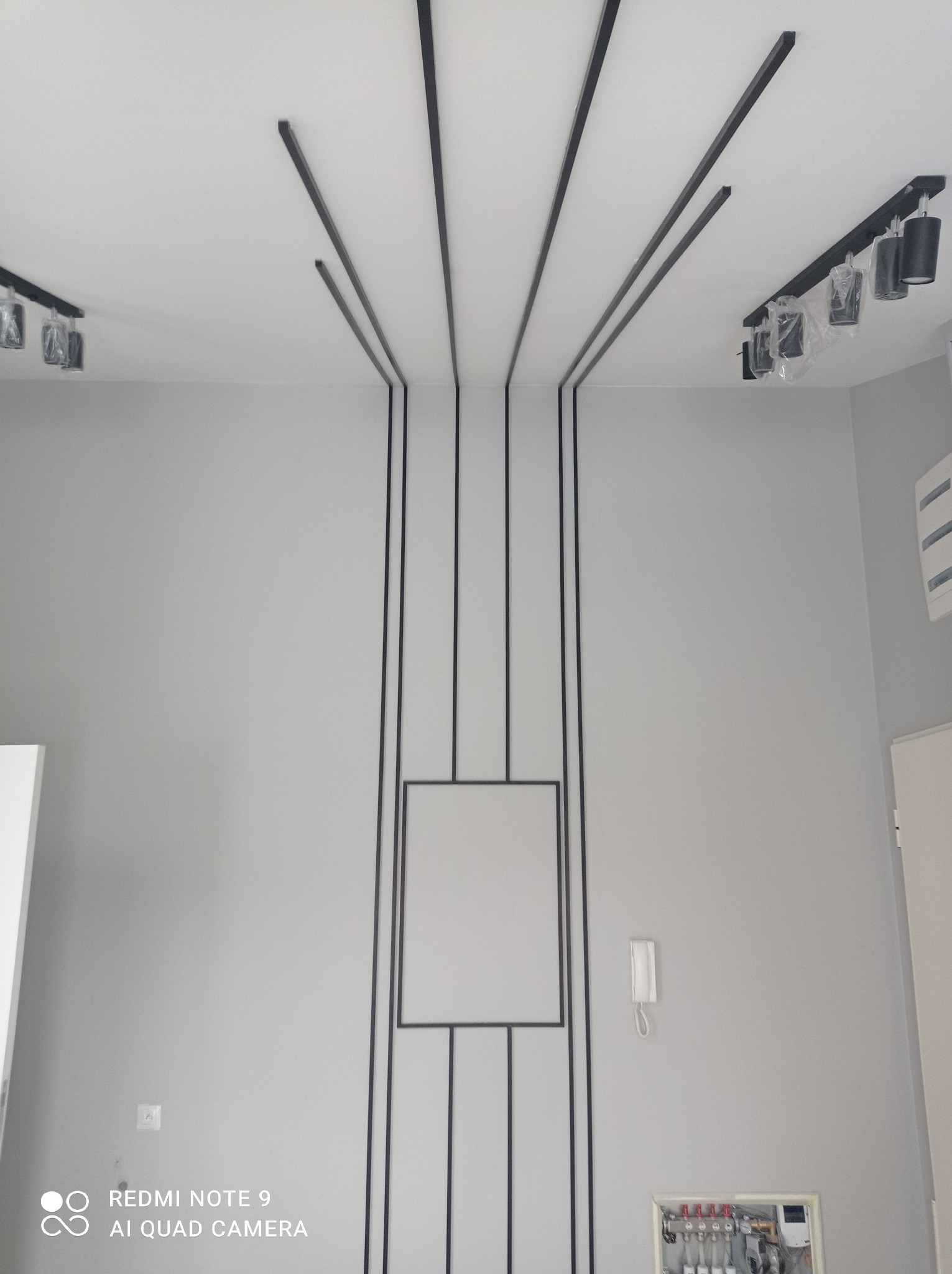 Szara ściana z dekoracyjnymi czarnymi listwami tworzącymi geometryczny wzór, oświetlenie sufitowe z czarnymi reflektorami, widoczna skrzynka z instalacją hydrauliczną.