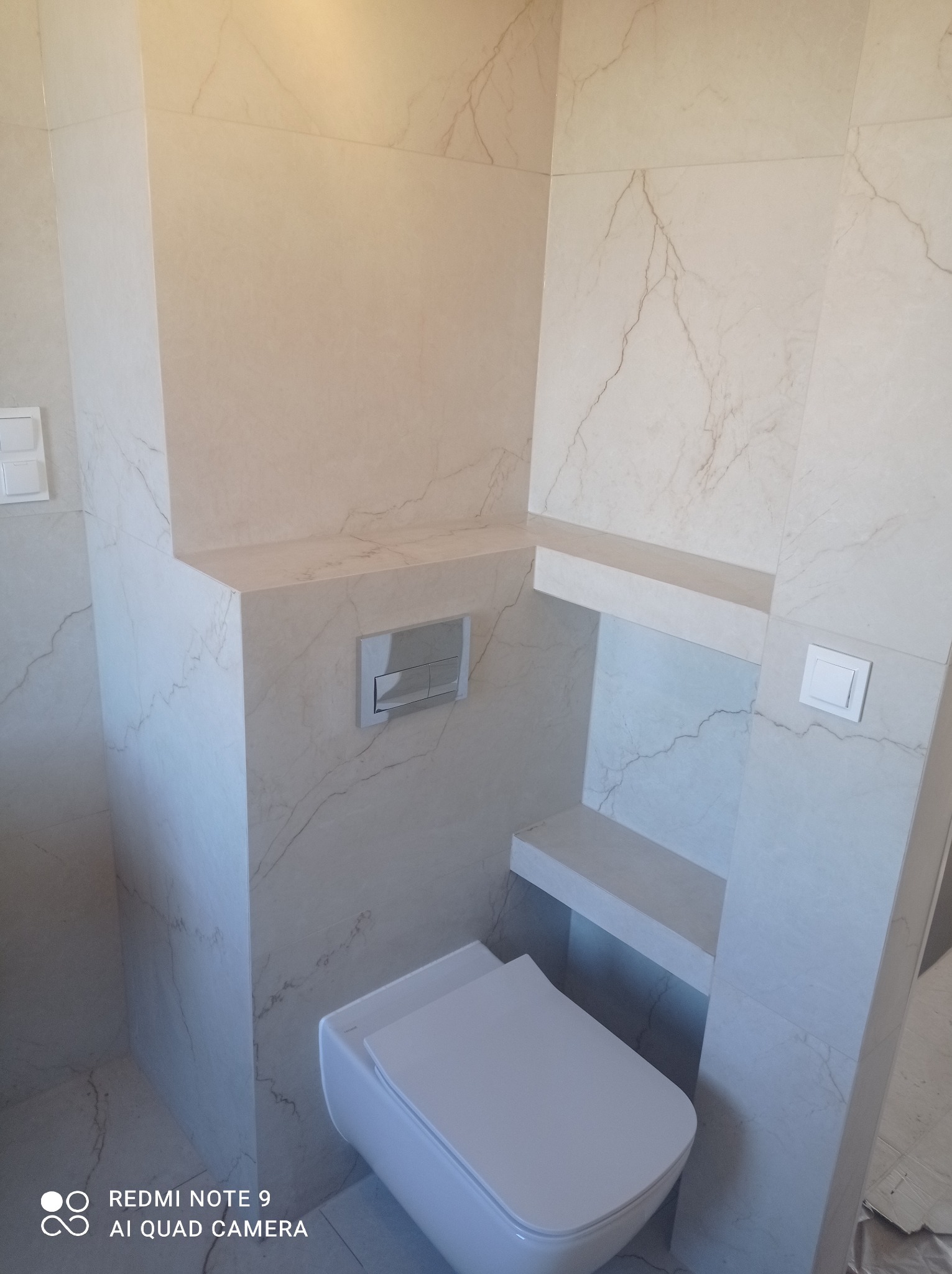 Wykończona toaleta w łazience z płytkami imitującymi marmur, zabudowa stelaża podtynkowego z półkami i przyciskiem spłukującym.