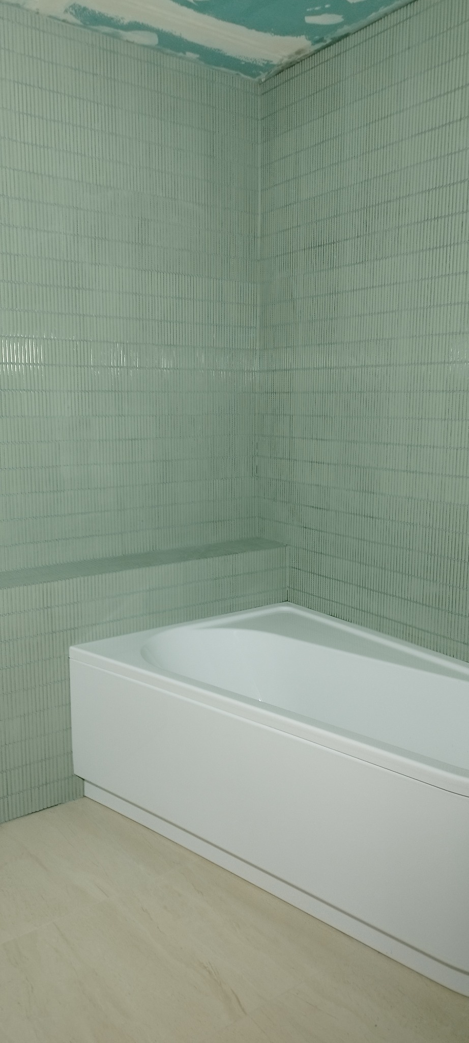 Biała wanna obudowana płytkami w łazience. Ściany wyłożone pionowymi, zielonymi płytkami. Na ścianie półka. Podłoga jasna, imitująca marmur.