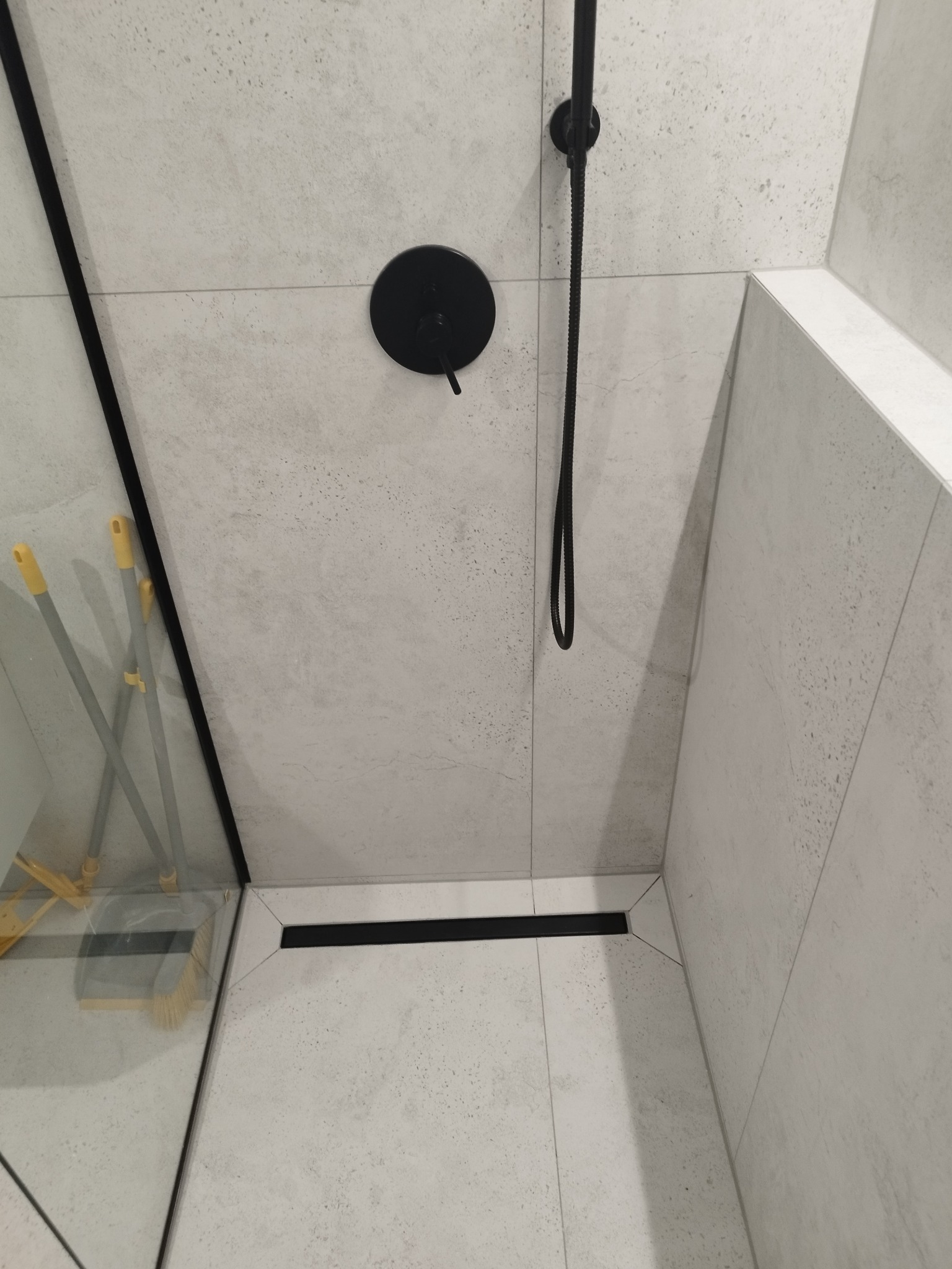 Nowoczesna kabina prysznicowa z szarymi płytkami, czarną armaturą i odpływem liniowym. Widoczne detale wykończenia i czystość.