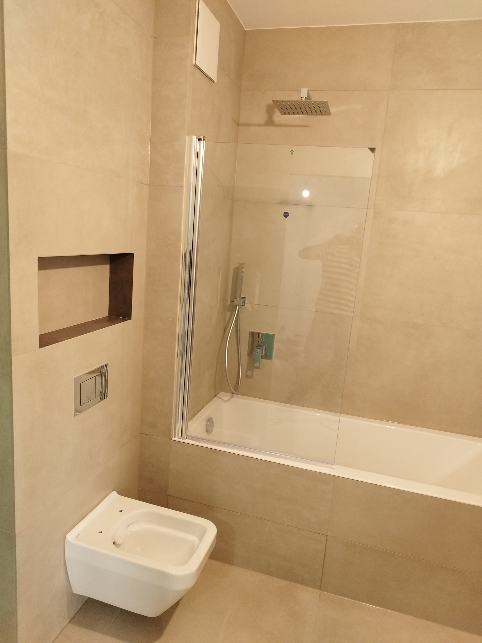 Beżowa łazienka z kabiną prysznicową, bidetem i wnęką w ścianie. Widoczne płytki w odcieniach beżu, szklana kabina i biała armatura. Minimalistyczny styl.