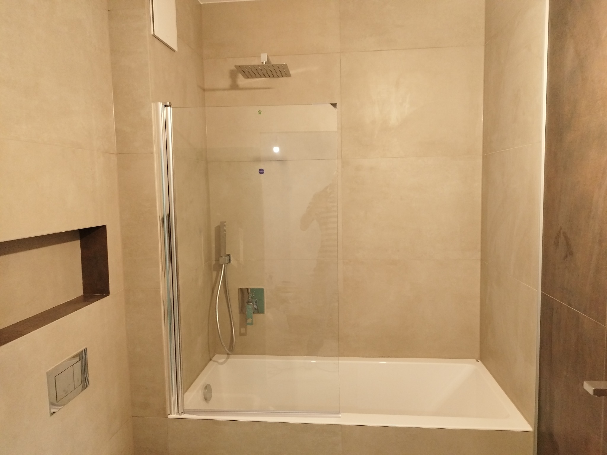 Beżowa łazienka z wanną i szklanym parawanem. Widoczna bateria prysznicowa, wnęka w ścianie i chromowany przycisk spłuczki. Minimalistyczny design.