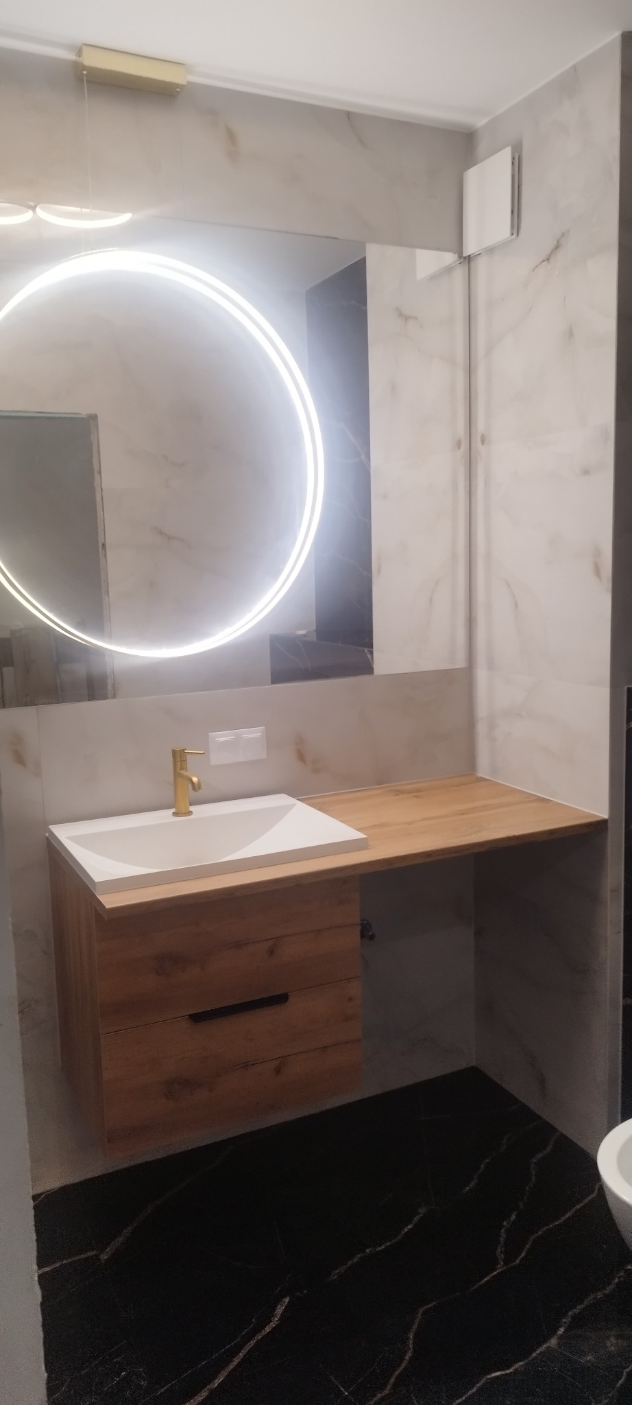 Nowoczesna łazienka z umywalką na drewnianej szafce, blatem i okrągłym lustrem LED. Marmurowe płytki na ścianach i czarna podłoga z białymi żyłami.