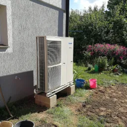 Zewnętrzna jednostka pompy ciepła Panasonic Inverter umieszczona na betonowych bloczkach przy szarej ścianie domu z widoczną instalacją wodną, obok ogród z kwiatami i narzędziami ogrodniczymi.