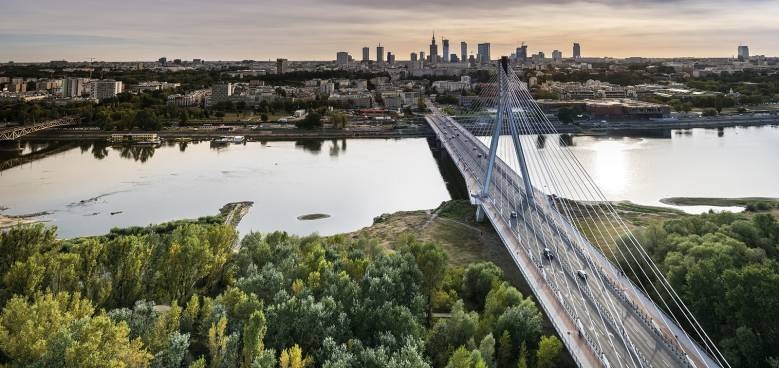 Most wantowy nad rzeką z panoramą miasta w tle, widok z góry na zieleń i wodę.