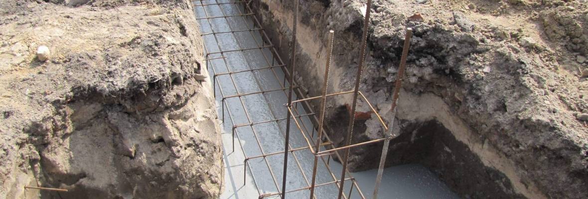 Wykop fundamentowy z zbrojeniem wypełniony świeżym betonem, widoczne warstwy ziemi w wykopie.