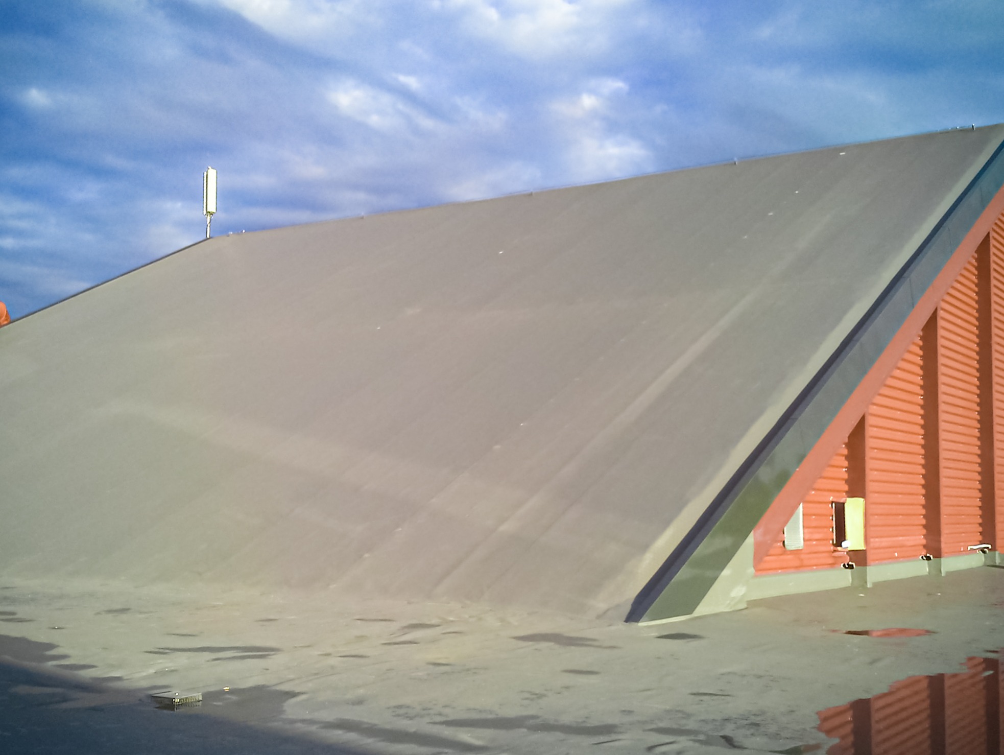 Szeroki, płaski dach pokryty szarą membraną z widoczną anteną na szczycie, przylegający do pomarańczowej, blaszanej konstrukcji z szarym obramowaniem, na tle pochmurnego nieba.