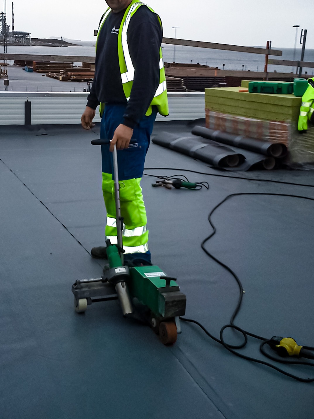 Pracownik w odblaskowej kamizelce używa zgrzewarki do papy na płaskim dachu, w tle widoczne rolki papy i materiały izolacyjne, a także fragment nabrzeża.