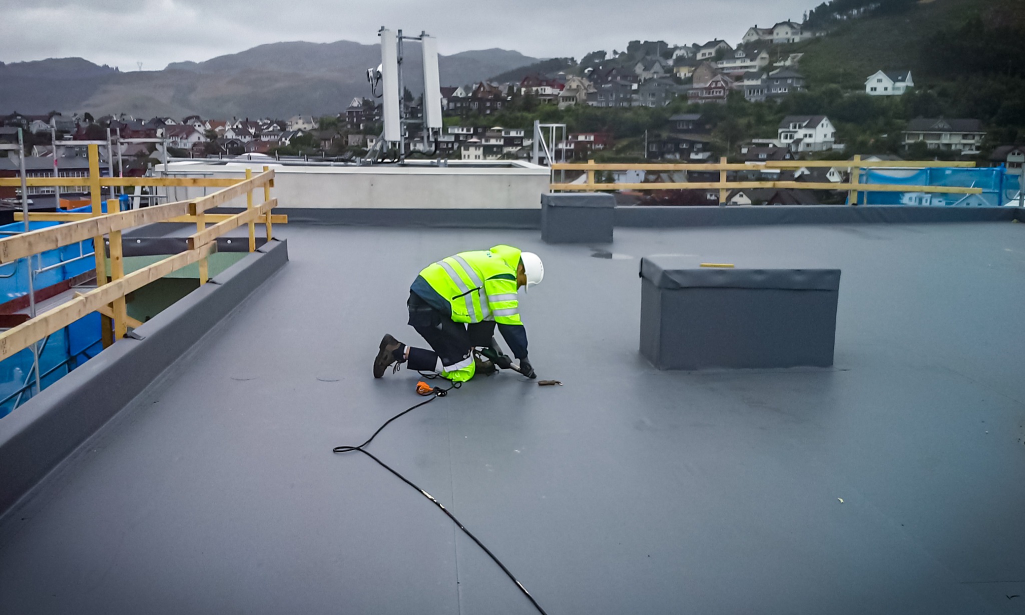 Pracownik w odblaskowej kurtce i kasku, na kolanach, używa narzędzia na płaskim, szarym dachu z widocznymi elementami wentylacyjnymi w kształcie prostopadłościanów; w tle zabudowa miejska...