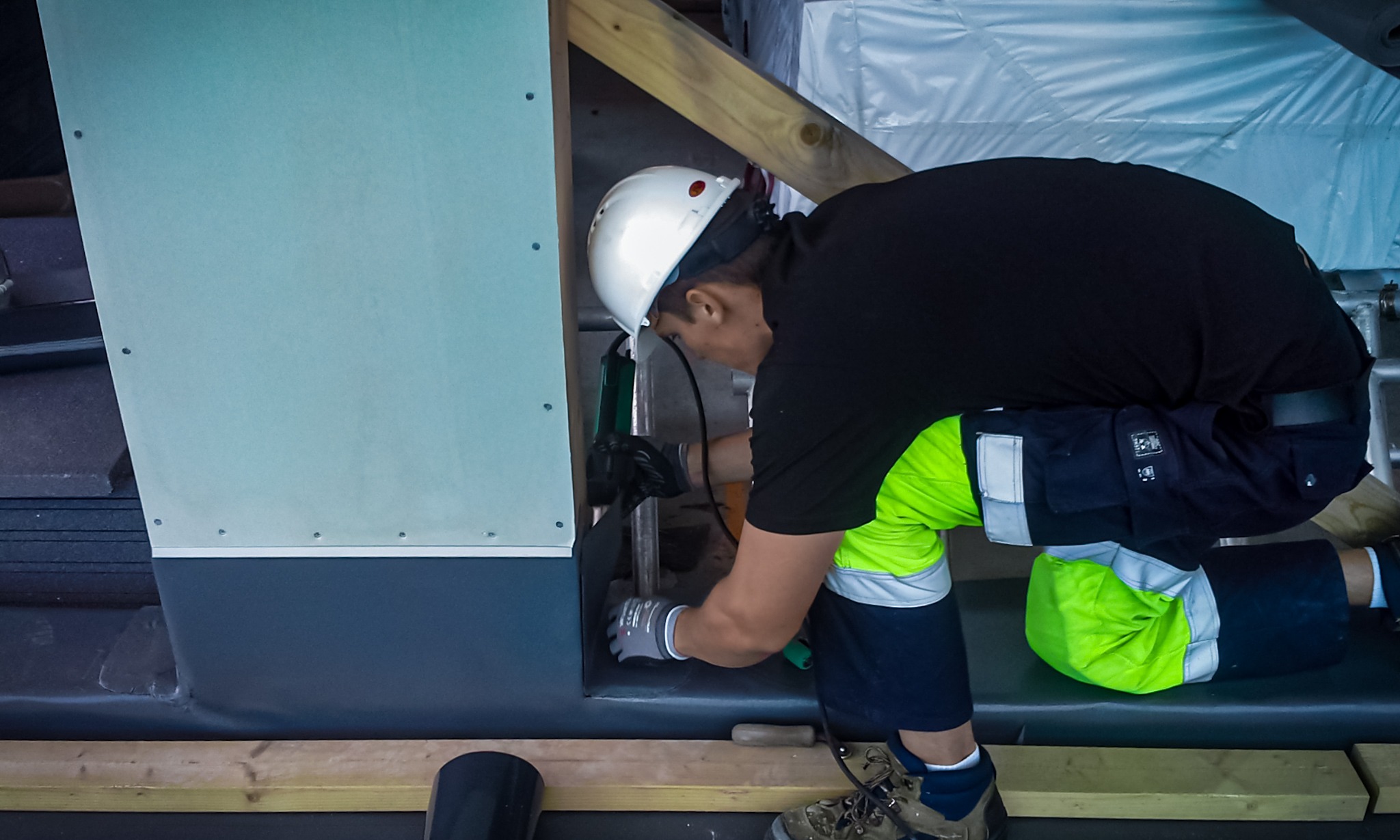 Pracownik w kasku i odzieży roboczej, z użyciem narzędzia, montuje izolację na fundamencie budynku, widoczne płyty izolacyjne i drewniane elementy konstrukcji.