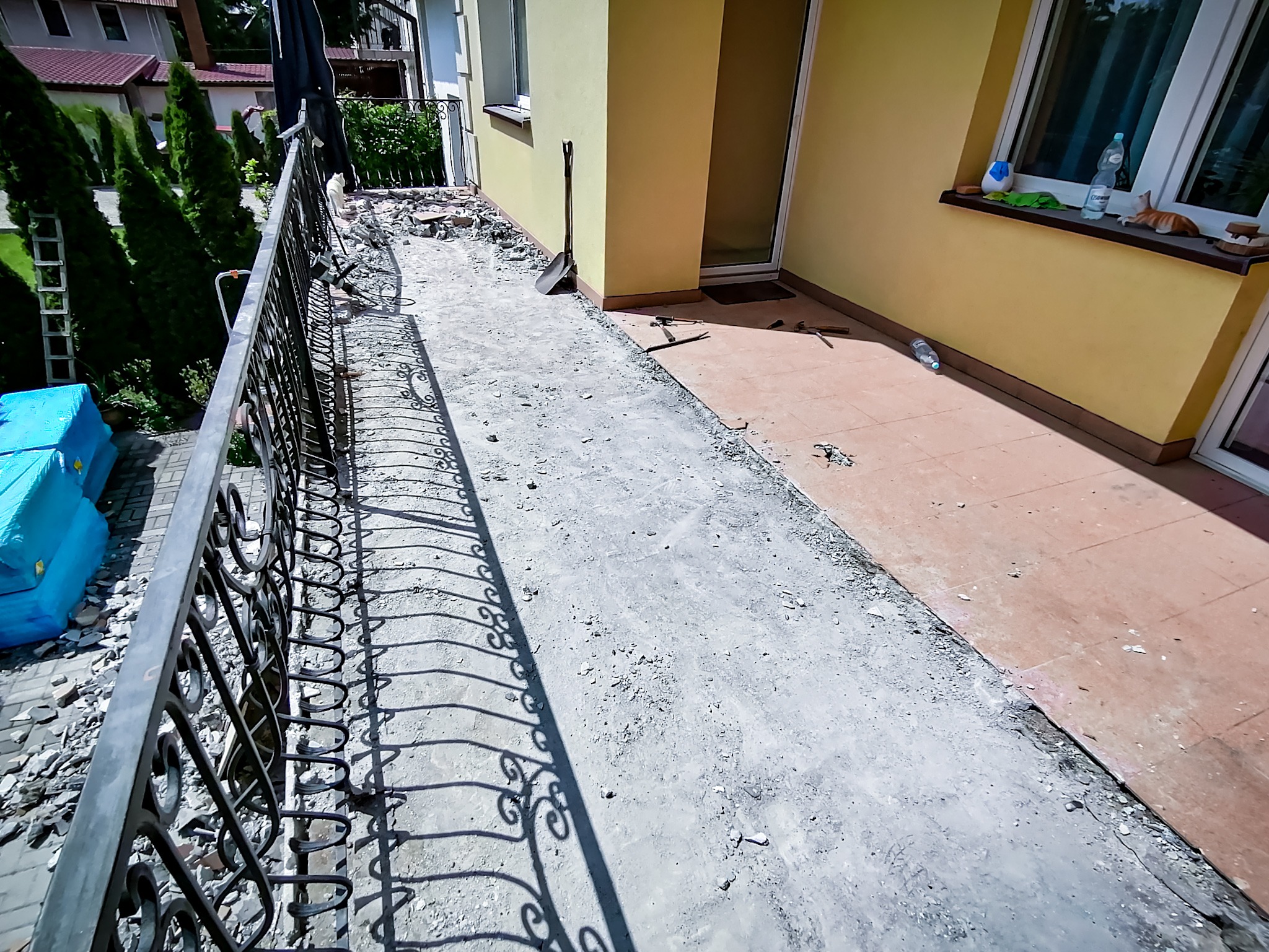 Przygotowanie podłoża na balkonie z metalową balustradą, widoczne narzędzia i resztki gruzu, żółta elewacja budynku w tle.