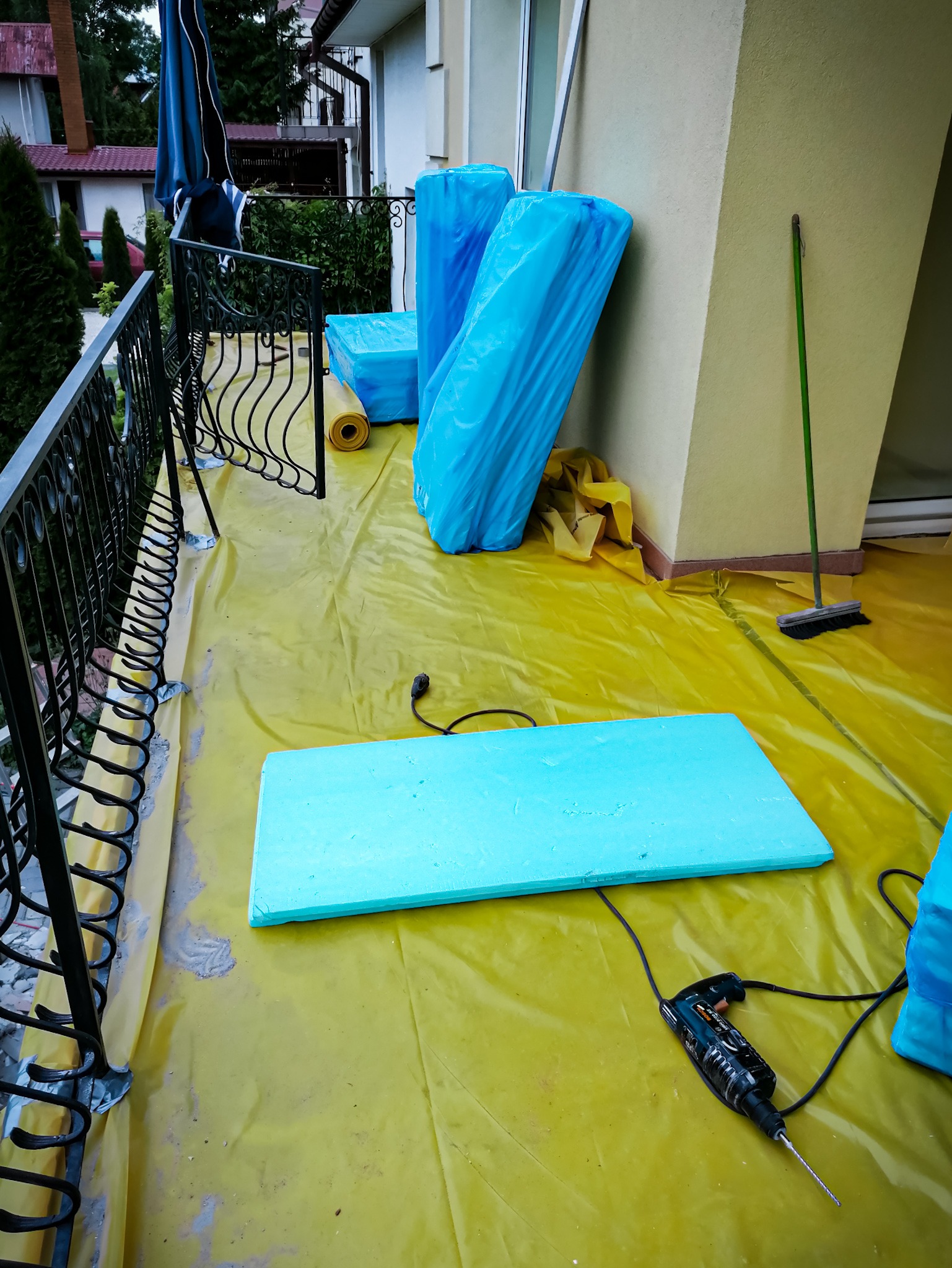 Przygotowanie do prac izolacyjnych na balkonie: styropian w niebieskiej folii, płyta styropianowa, wiertarka na żółtej folii ochronnej, widok z góry.