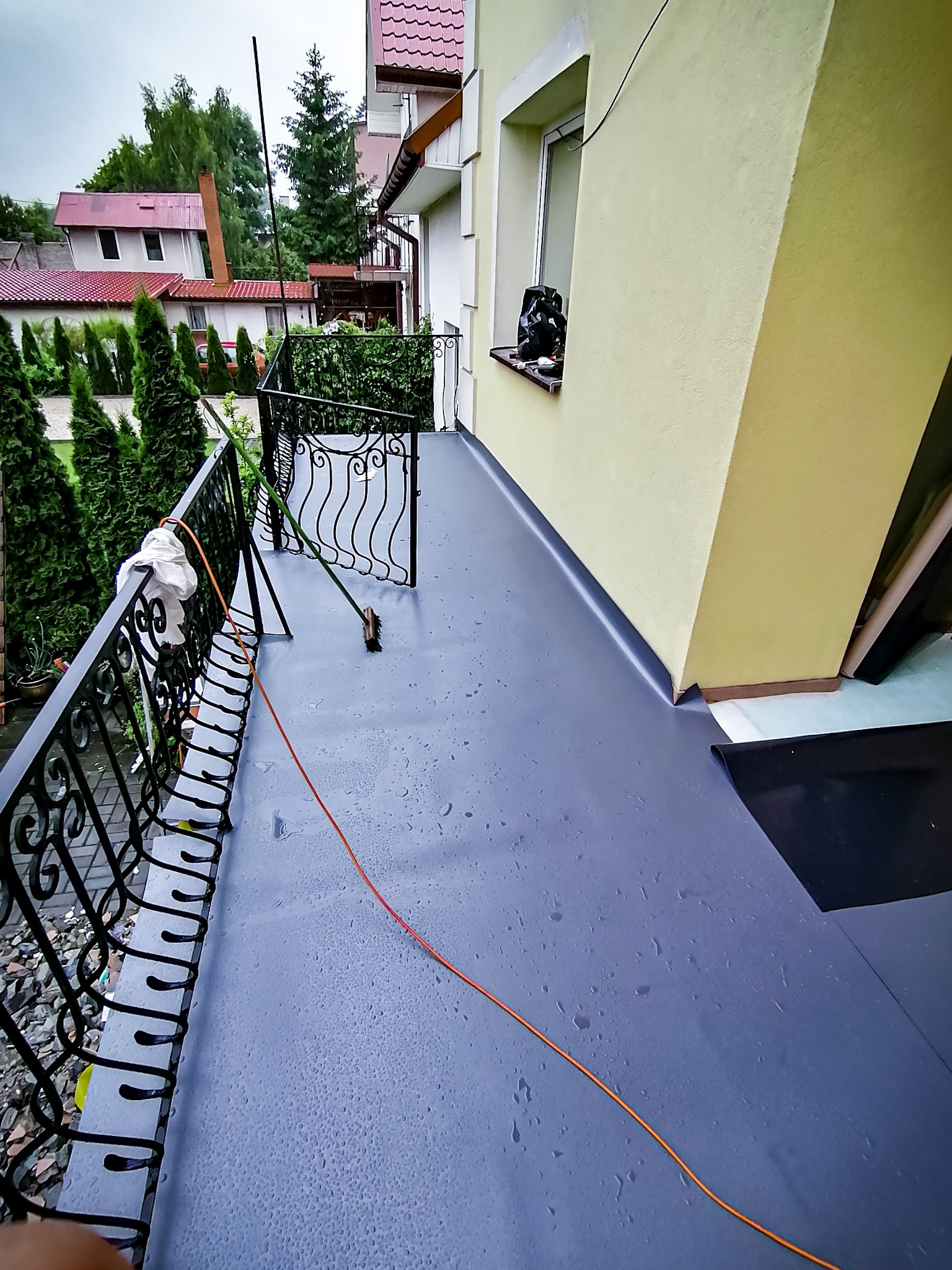 Szary, płaski dach balkonu po hydroizolacji, widoczne krople wody, czarna balustrada z ozdobnymi elementami, żółta ściana budynku i fragment okna, w tle zieleń drzew i zabudowania.