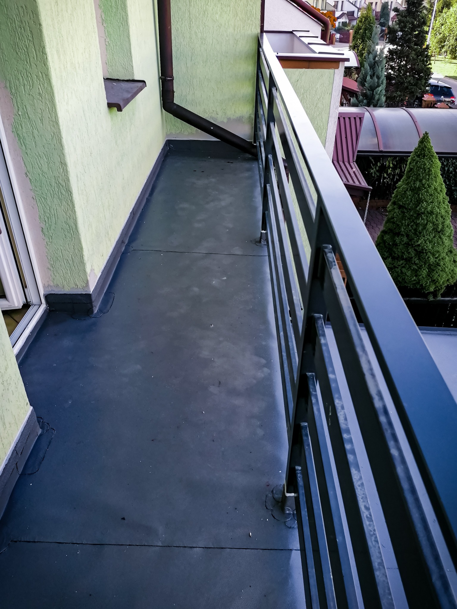 Wykończony balkon z nową, szarą hydroizolacją, widok na metalową barierkę i otoczenie domu z zielonymi drzewami.
