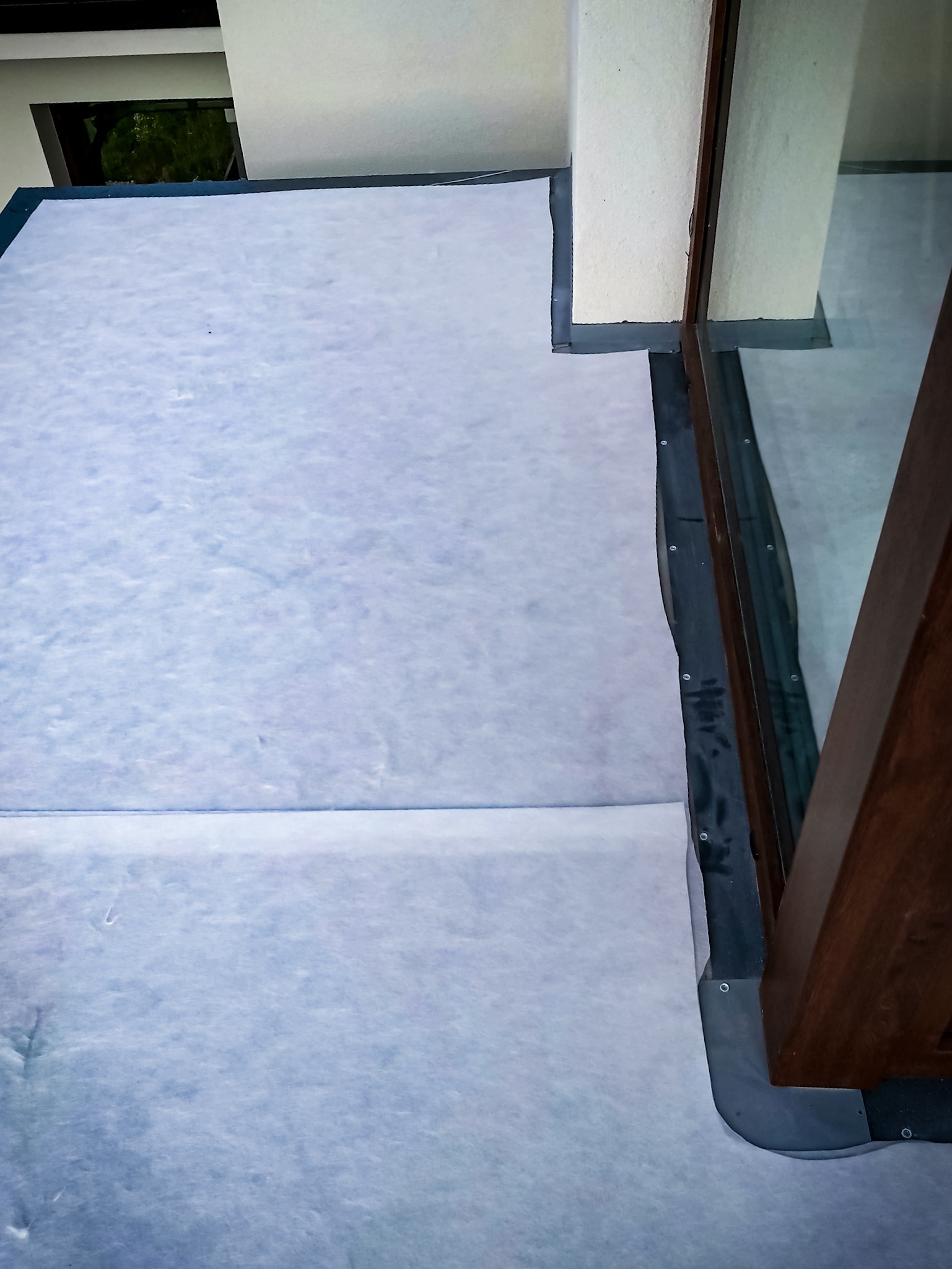 Biała membrana dachowa rozłożona na dachu, widoczne detale mocowania przy oknie dachowym, szara blacha obróbki blacharskiej.