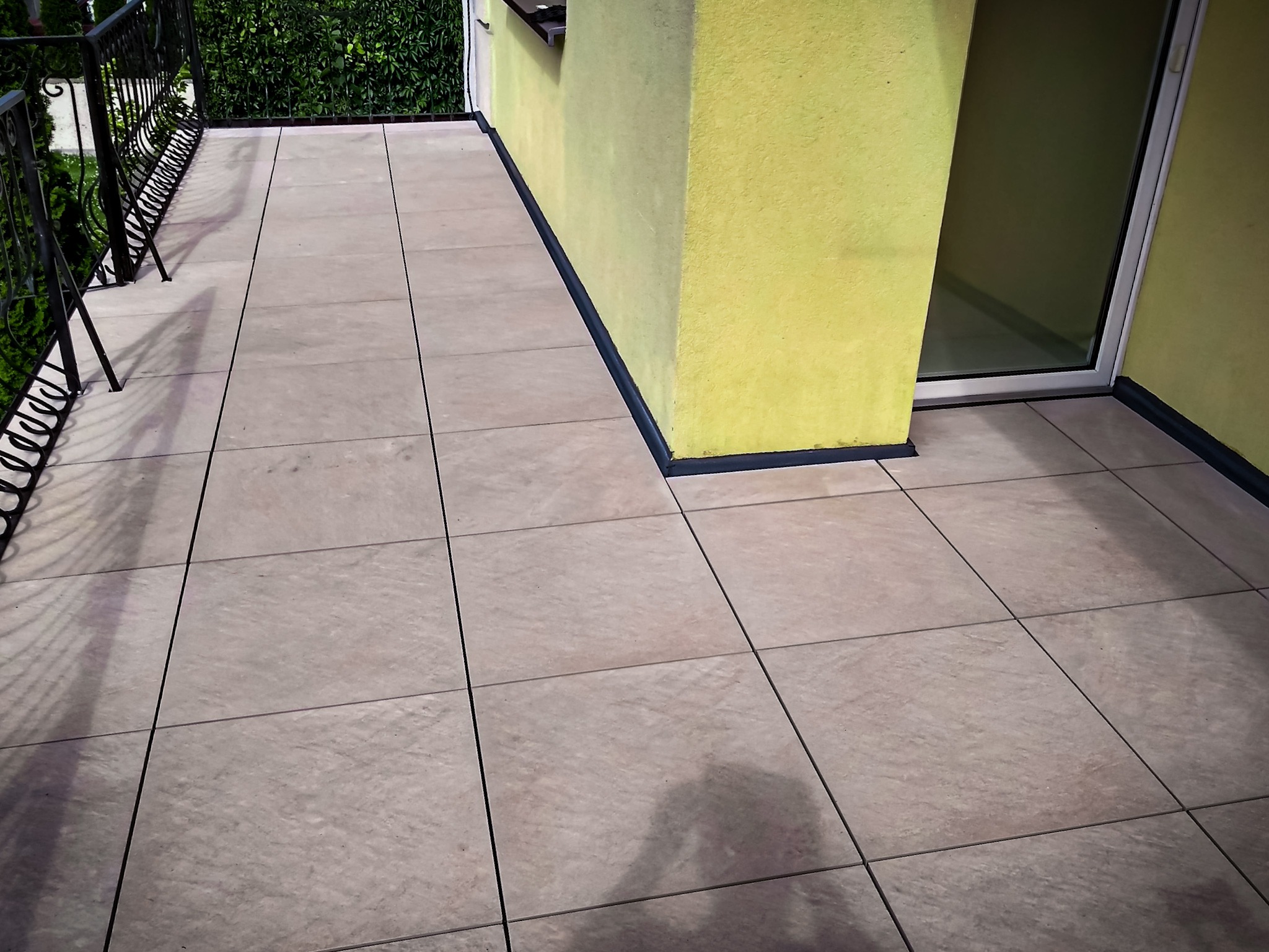 Wyłożony jasnymi płytkami taras z metalową balustradą i żółtą ścianą budynku z oknem balkonowym.