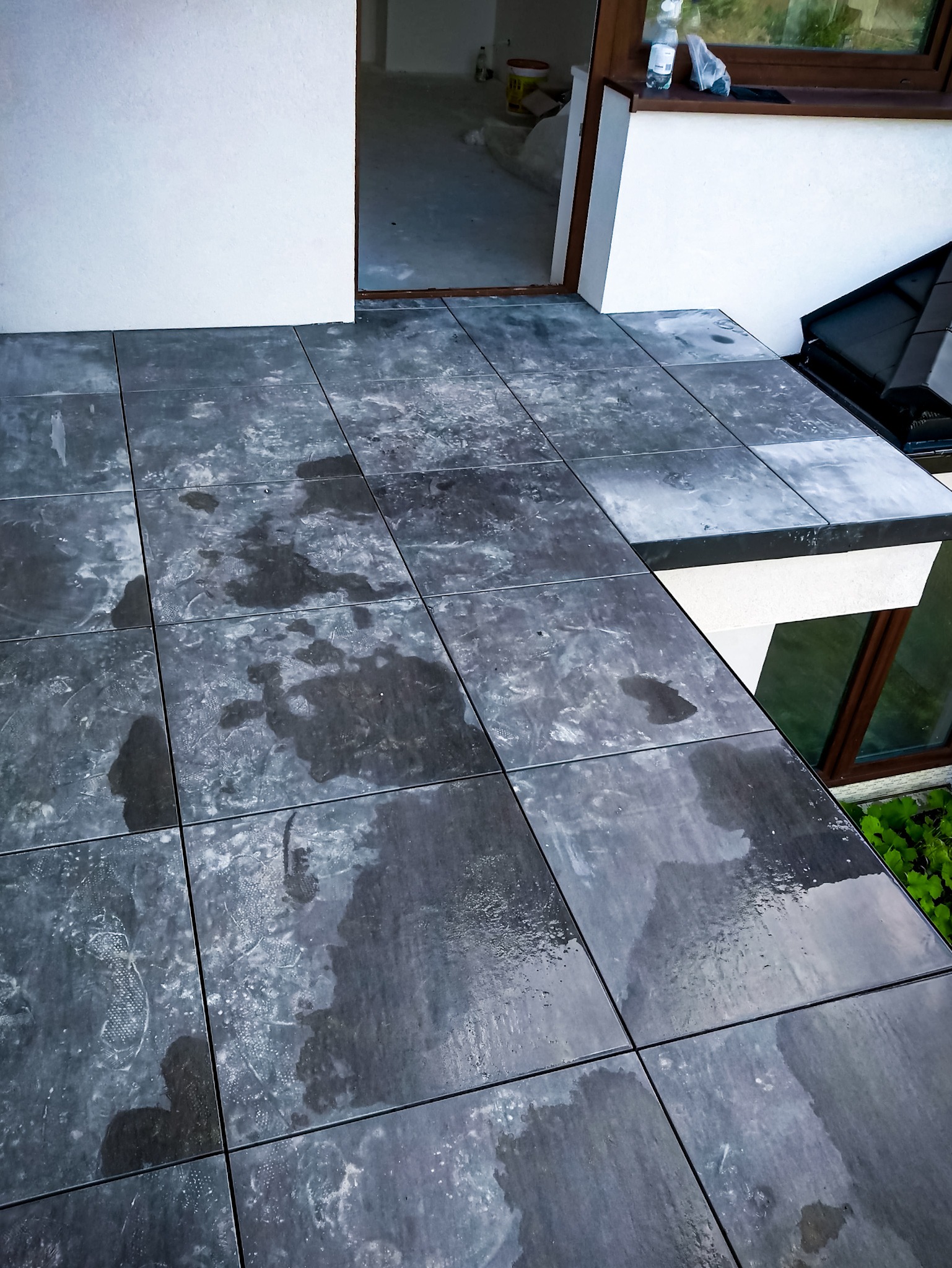 Szare płytki na tarasie z widocznymi śladami wody i odciskami butów, prowadzące do otwartego wejścia do budynku.