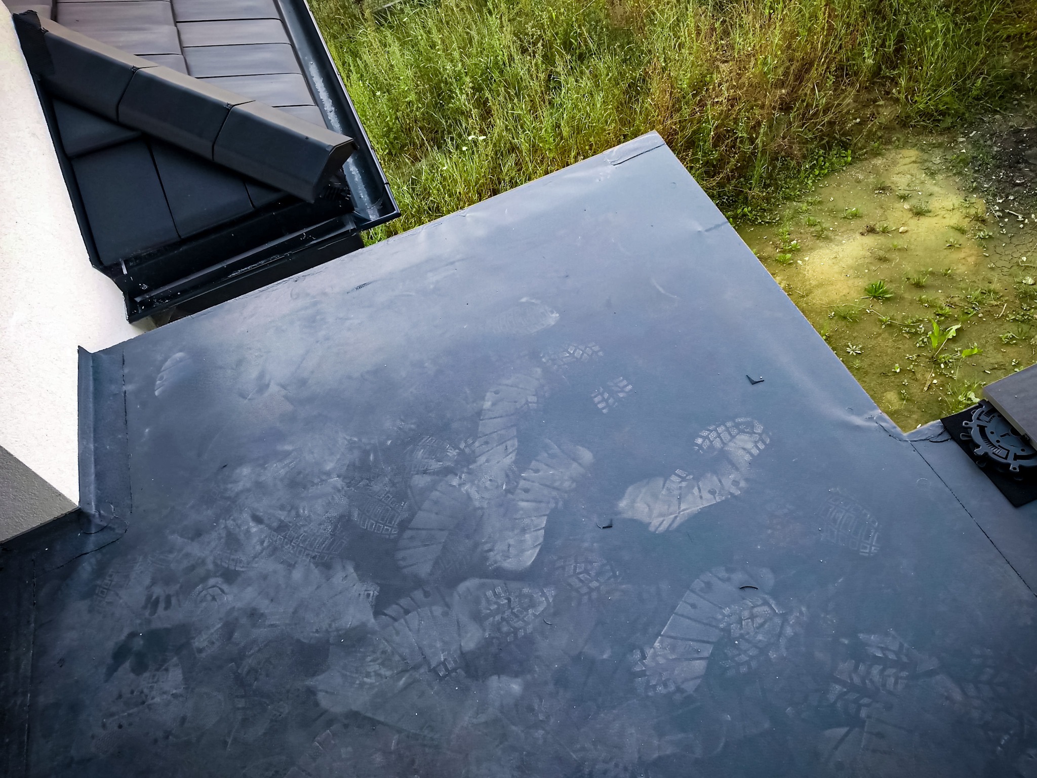 Widok z góry na fragment płaskiego dachu pokrytego papą bitumiczną z widocznymi odciskami butów, obok rynna i fragment dachu z dachówki ceramicznej oraz zarośnięte pole w tle.