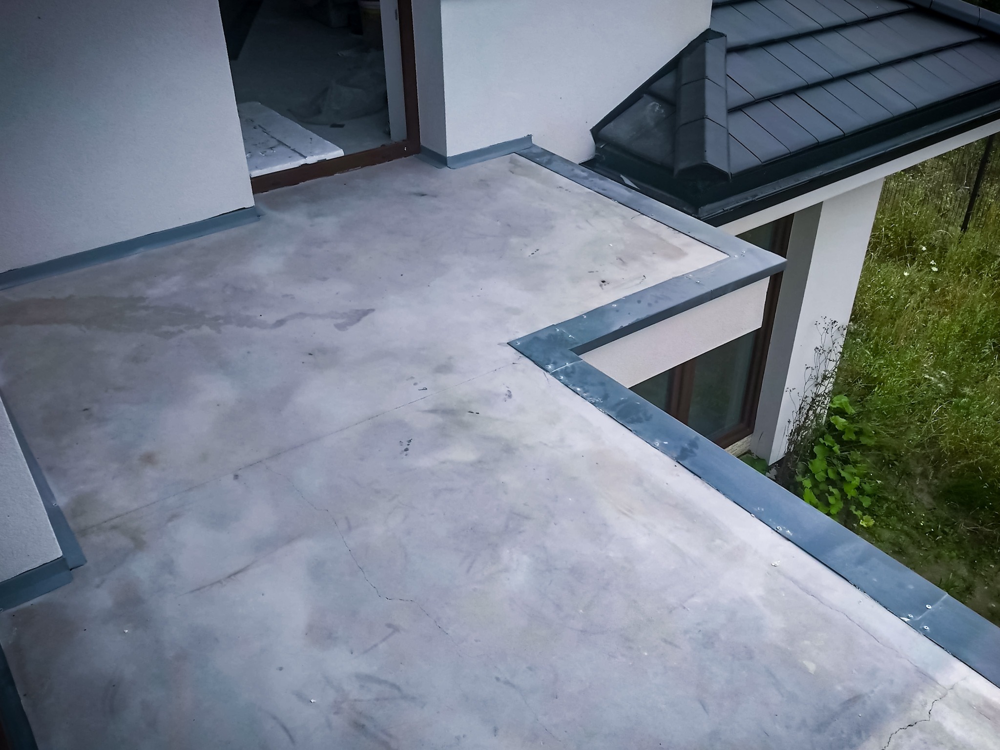 Wykończony taras na dachu z szarą hydroizolacją i widokiem na dach z czarną dachówką oraz zieloną roślinność.