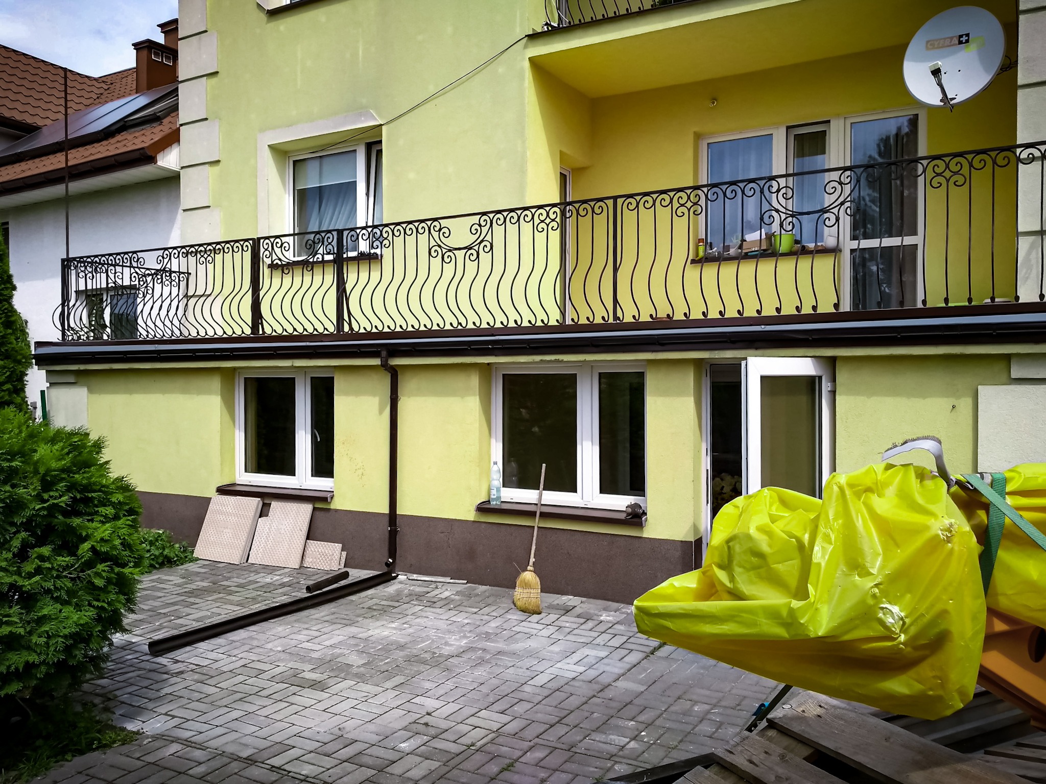 Dwupiętrowy dom z balkonem i widocznymi elementami przygotowania do prac budowlanych, w tym zabezpieczona folią żółtą sterta materiałów i stojąca miotła.
