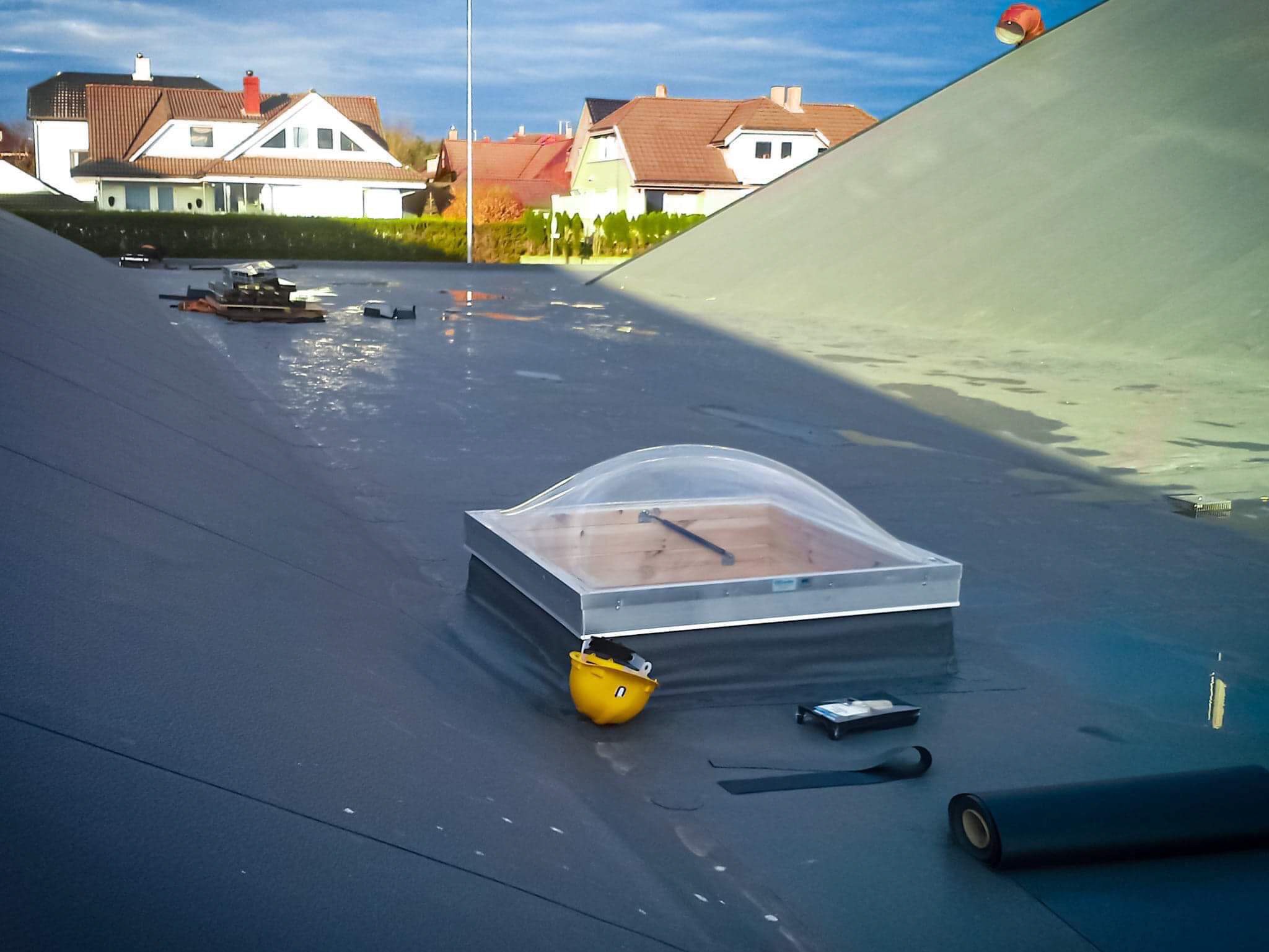Membrana dachowa z zamontowanym świetlikiem kopułkowym, żółty kask obok, narzędzia dekarskie i rolka papy na płaskim dachu z widokiem na zabudowę jednorodzinną.