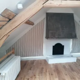 Poddasze po remoncie: drewniane belki stropowe, ściany w pionowe pasy, biały grzejnik żeberkowy, drewniana podłoga, biały kominek z czarnym wkładem i szarym okapem.