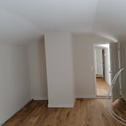 Poddasze po remoncie: Świeżo pomalowane białe ściany, jasna drewniana podłoga, otwarte drzwi do kolejnego pomieszczenia z widoczną podłogą. Białe listwy przypodłogowe.